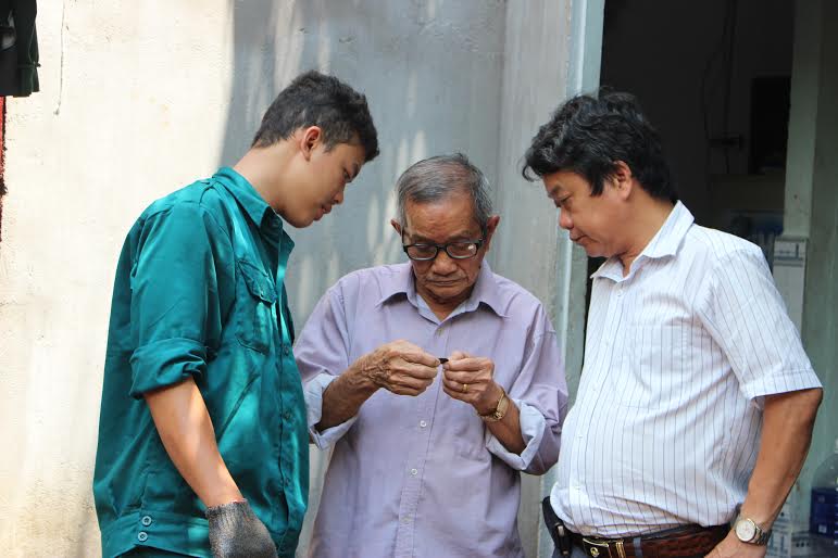 Ông Phan Văn Lai (giữa) và ông Hồ Văn Lộc, Phó Giám đốc Sở Lao động - thương binh và xã hội (bên phải) đang xem di vật là đồng tiền xu tìm thấy dưới hố chôn.